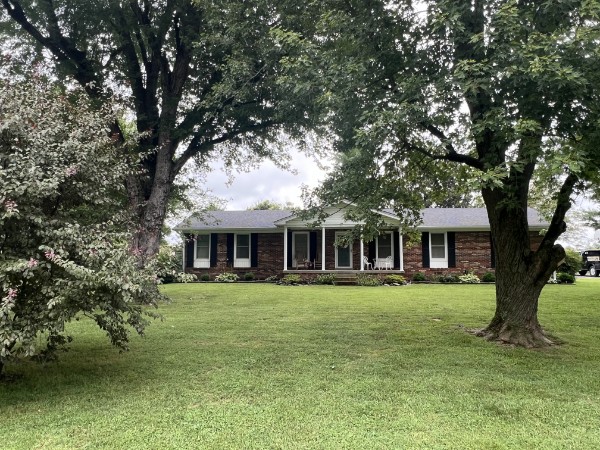 Residential real estate auction home — single‑story brick house with white shutters, central porch, and landscaped front yard framed by mature trees.